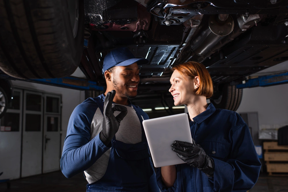 Smiling african american mechanic showing ok gesture near colleague with digital tablet under car in service